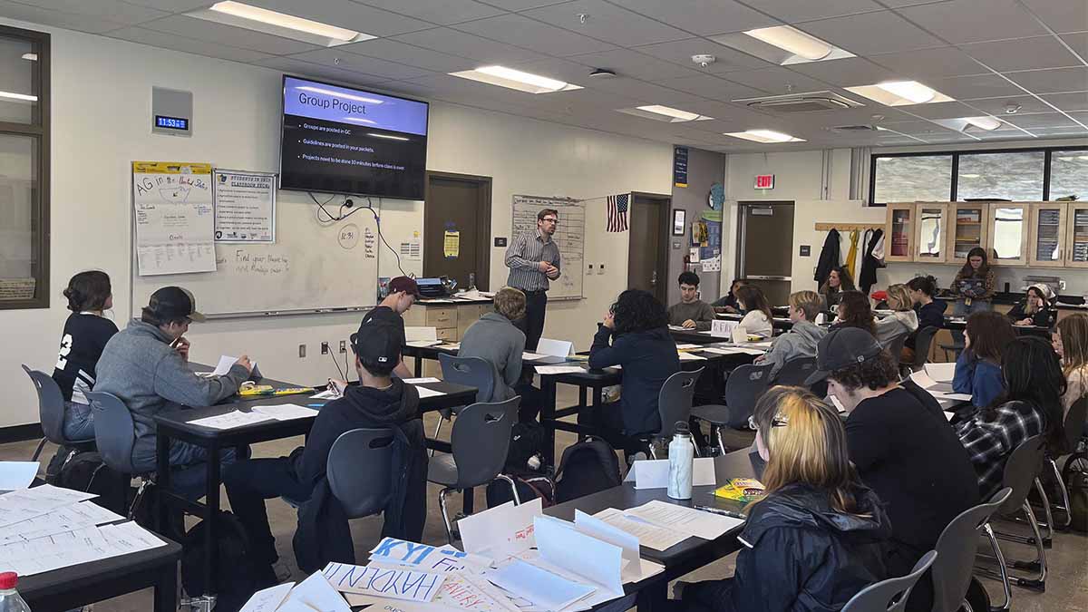 A male teacher stands in front of a classroom of high school students.