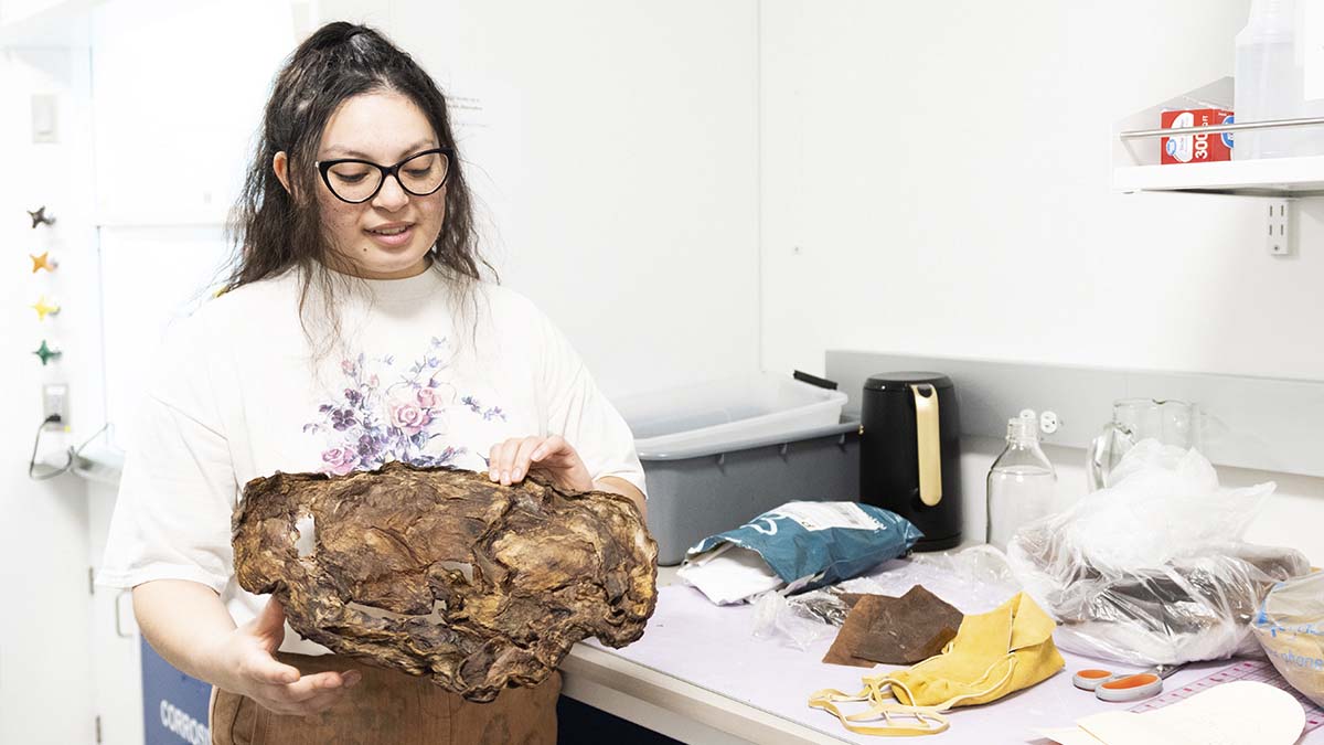 A woman holds a large, dried SCOBY in a lab next to a table with patterns, scissors and a leather moccasin.