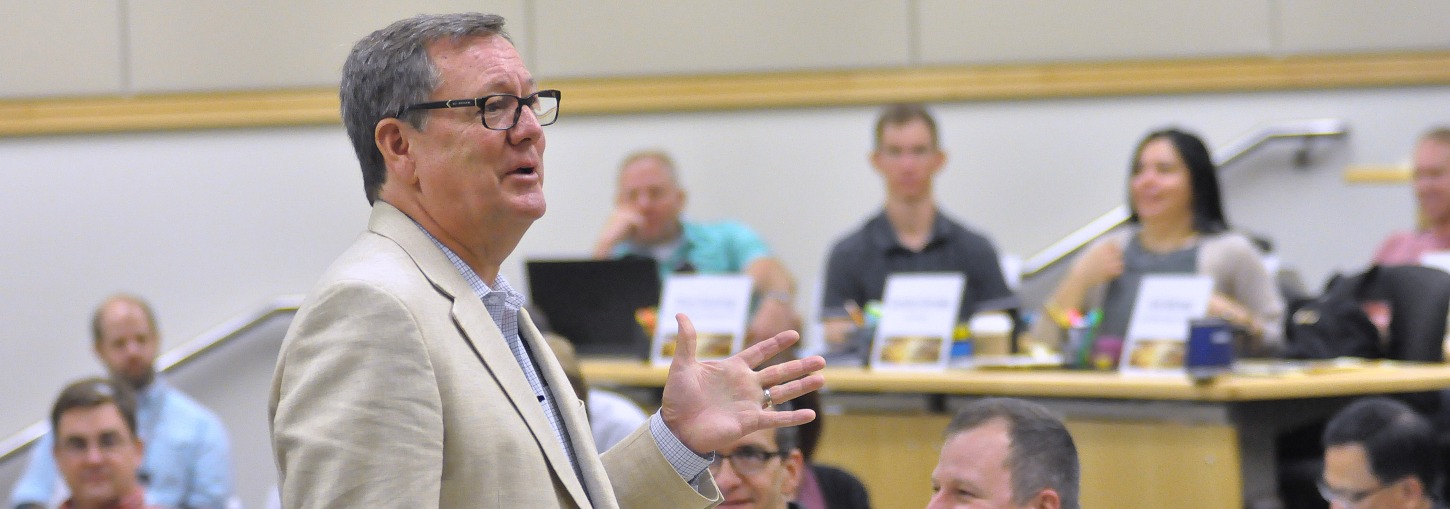 A man gestures while standing at the front of a classroom. Energy Executive Course participants seated in a modern classroom are visible out of focus in the background.