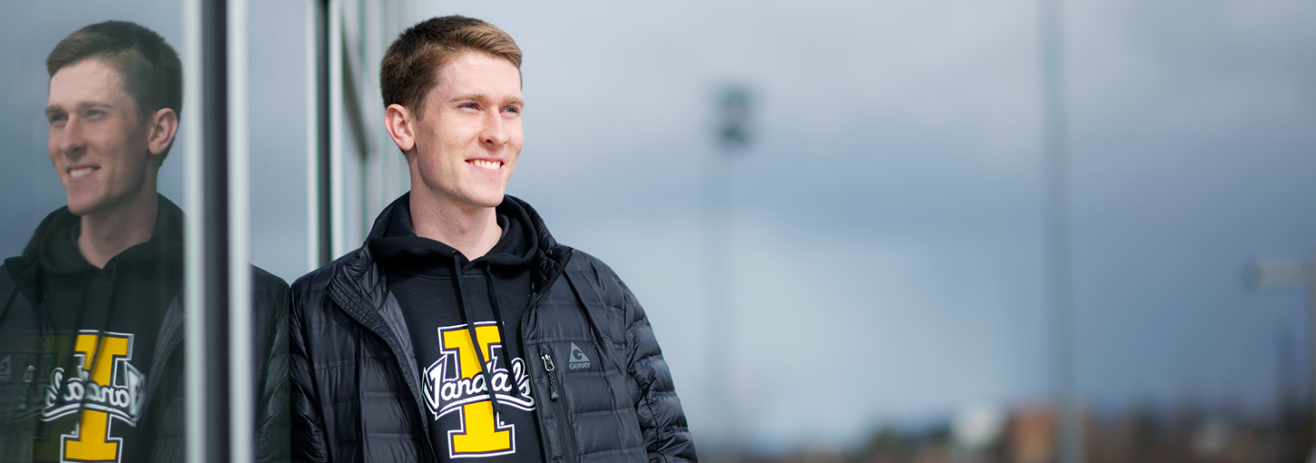 Young man wearing University of Idaho sweatshirt and black jacket leaning against a window at a sport complex.