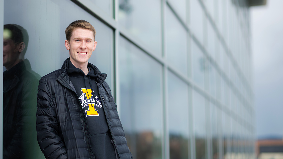 Young man wearing University of Idaho sweatshirt and black jacket leaning against a window at a sport complex.