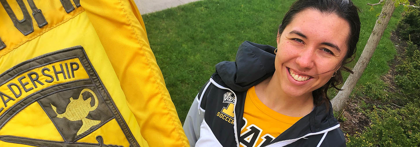 Woman wearing Vandal gear stands behind fluttering Army ROTC battalion guidon.