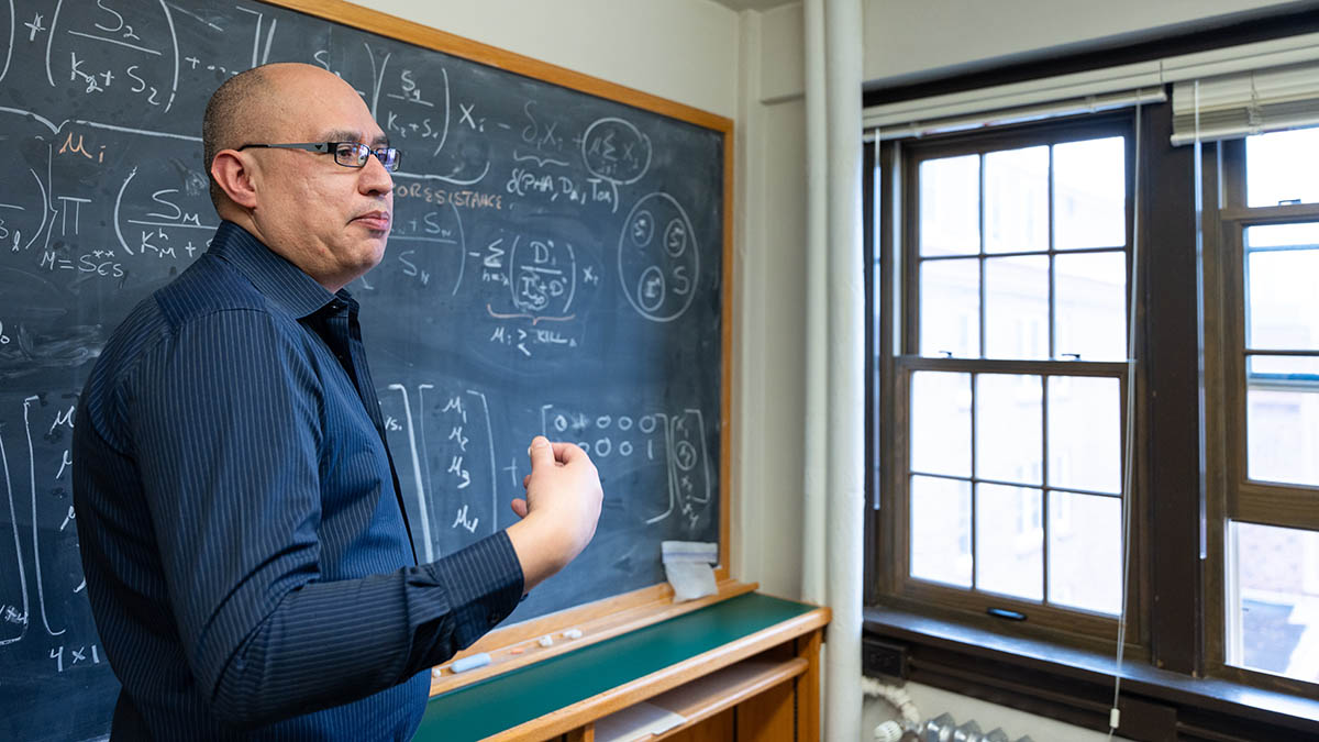 Man wearing glasses and blue shirt motions with his hand while standing in front of a mathematical formula on blackboard.