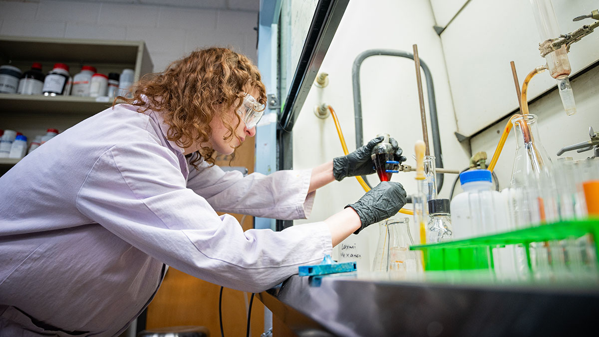 Woman in chemistry lab wearing safety goggles and lab coat examines a beaker.